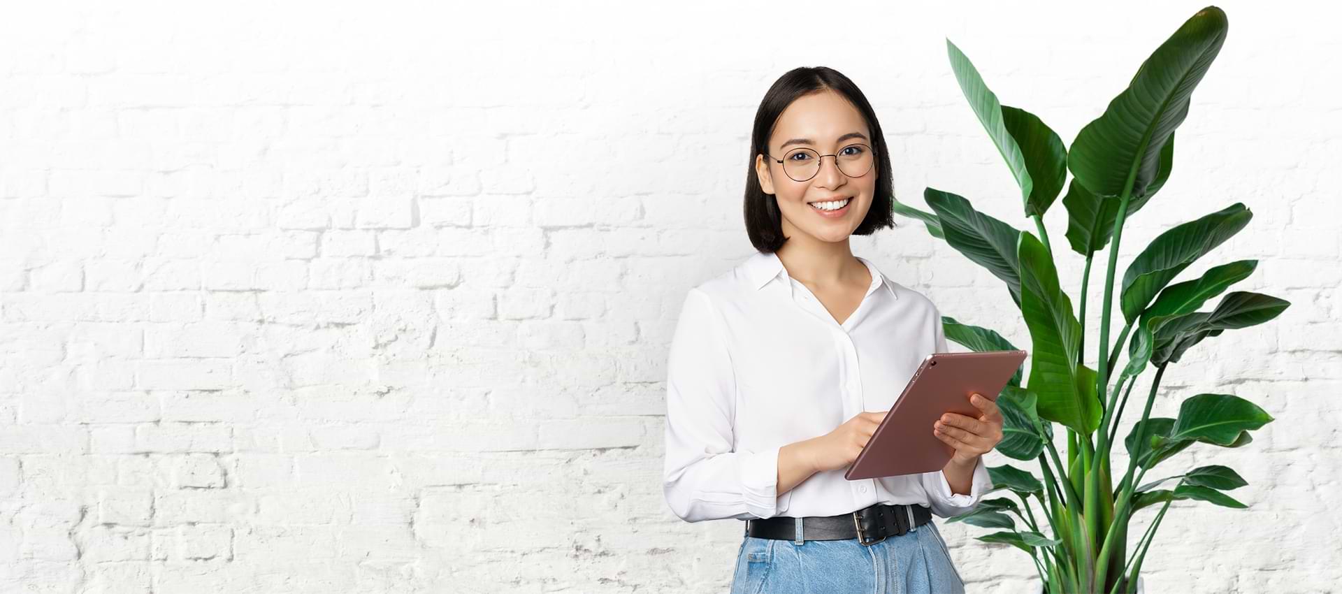 Woman holding a tablet with a white brick background standing next to a large plant